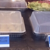 Labeled covered trays on a buffet table, including corn on the cob